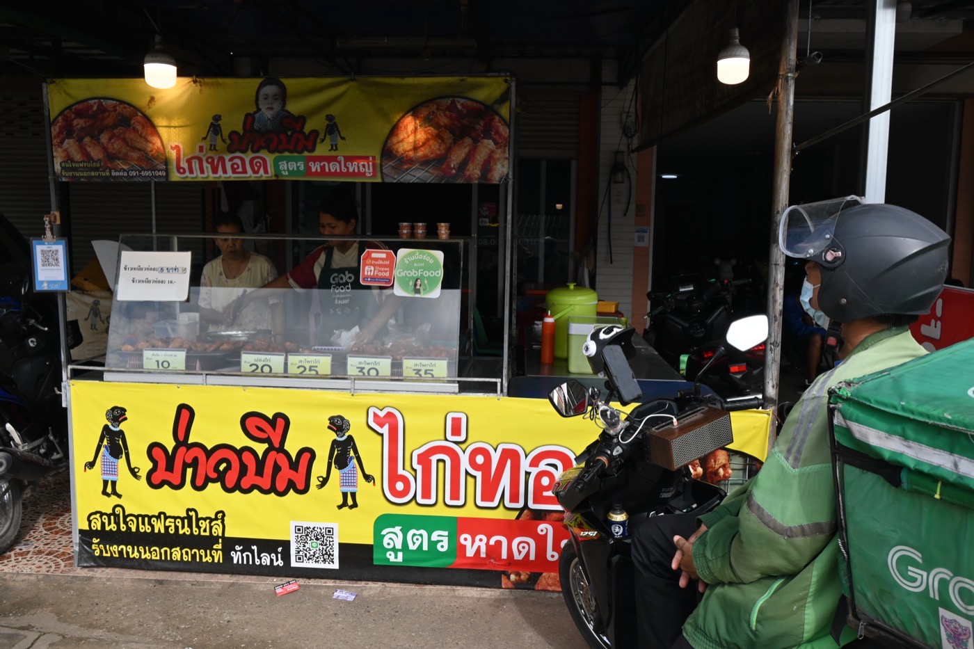 A GrabFood rider waits on his motorbike at a brightly painted street food stall